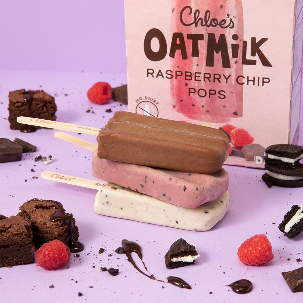 brownie batter, raspberry chip and cookies and cream pops stacked on a purple background with a raspberry chip box in the background and pieces of brownies, cookies, and raspberries around